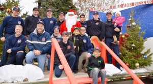 Caption:  Lockwood Fire Chief John Staley posing with Santa along with his crew and volunteers at Canary and Bluebird in Lockwood during one of the visits.  It takes a crew of about a dozen people to prep, help, take down and then move the Santa set from location to location.  (Photo by Jonathan McNiven)    